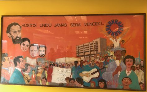 Mural featuring Hostos' main building in the background, surrounded by people apparently celebrating, playing music, and holding protest signs. The head of Eugenio María de Hostos floats in the sky. Text at top says "Hostos Unido Jamas Sera Vencido."